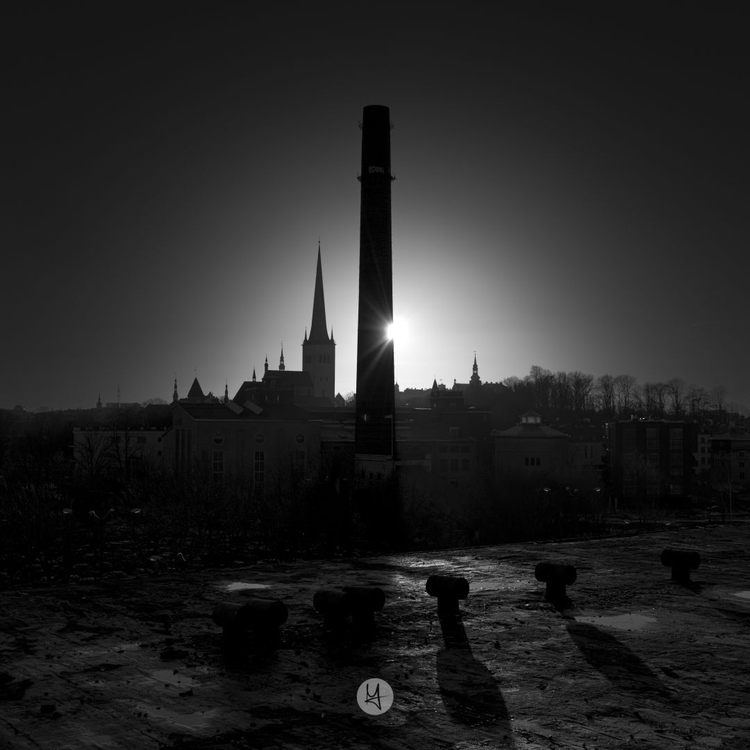 Square black and white photo, view of the old town from the top of the Linnahall at sunset. The sun is peaking behind a huge chimney surrounded by the tops of churches from the old town.