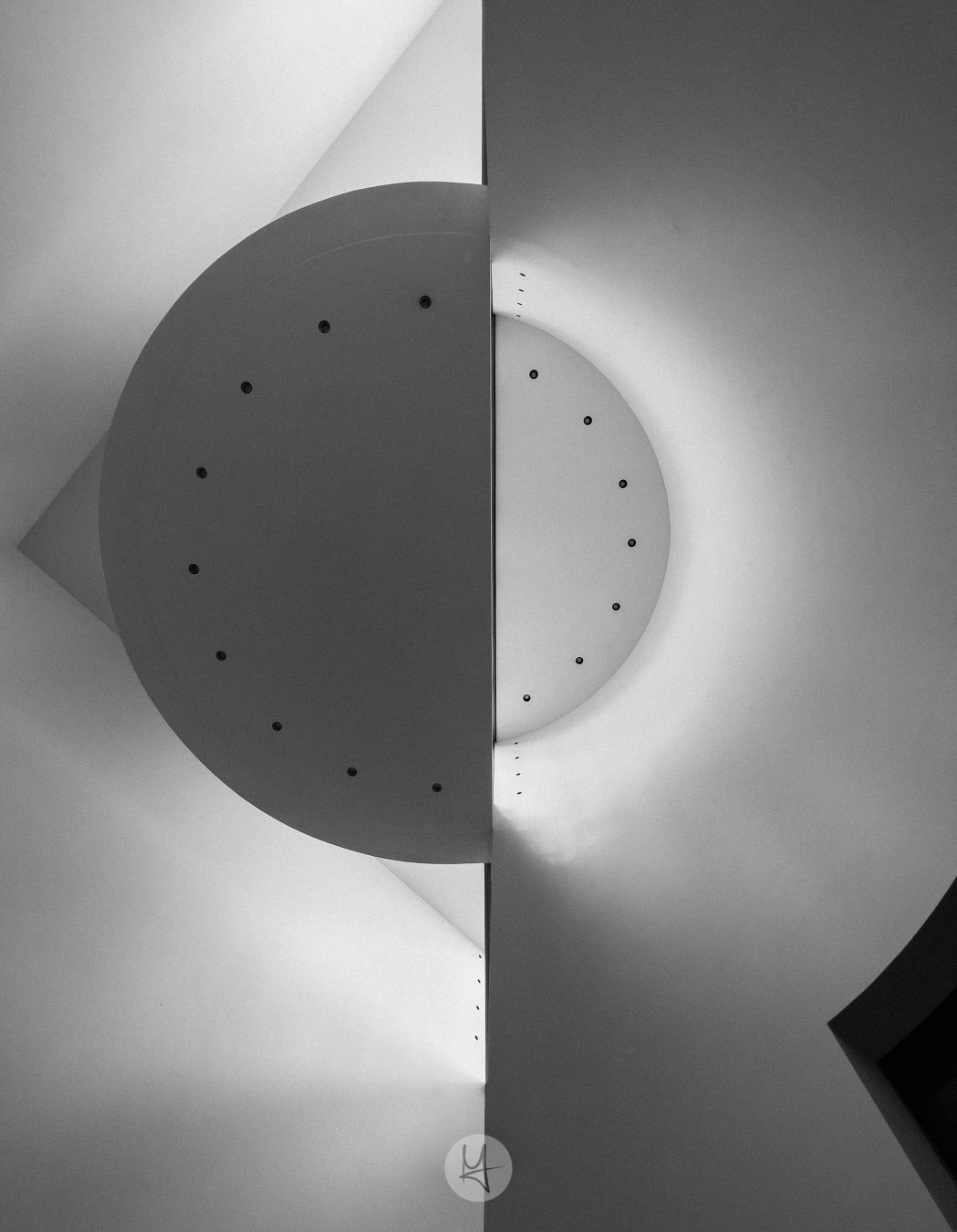 A black and white photograph shows a section of a modern building's interior.  The main focus is a large, semi-circular ceiling element split down the middle by a vertical wall. Both halves of the semi-circle have small, evenly spaced, dark circular fixtures along their edges. The walls and ceiling are smooth and appear to be made of a light-colored material.  A bright light source illuminates one side of the semi-circle, casting a shadow on the other. The overall composition is geometric and abstract.