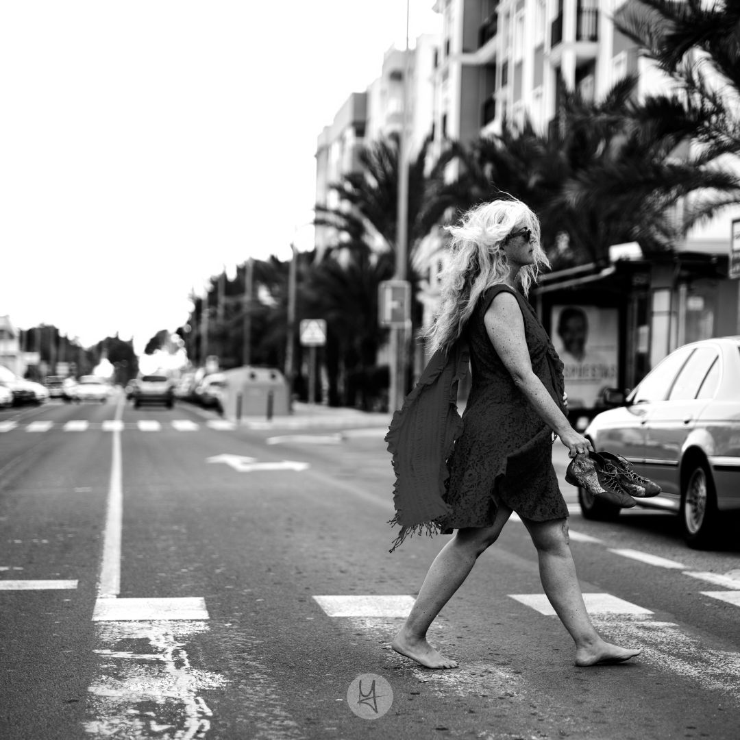 A black and white photo of a barefoot woman with long, windblown hair, crossing a street in a dark-colored dress. She is carrying a pair of shoes in her right hand.  The street is relatively empty with a few cars in the background, and palm trees line the street. Buildings are visible in the distance.  The image is taken at a slight angle, showing the woman from the side.