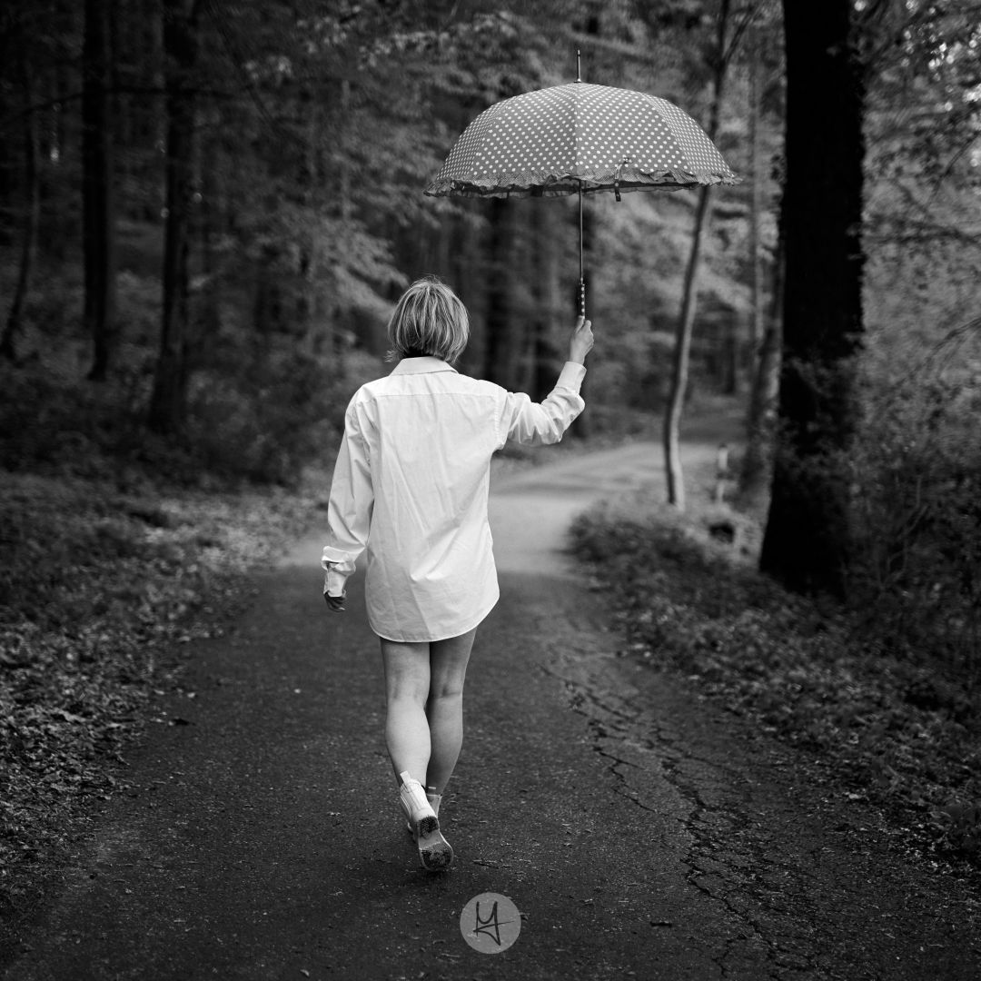 A black and white photo shows a woman with short hair, wearing a bright rain coat and rubber boots, walking away from the camera down a path in a wooded area. The person is holding a polka-dotted umbrella. The path is paved and the trees along the sides are dark and densely packed.